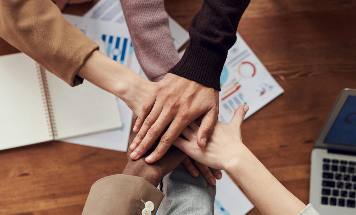 People put their hands together above a wooden table with data sheets and a laptop on it