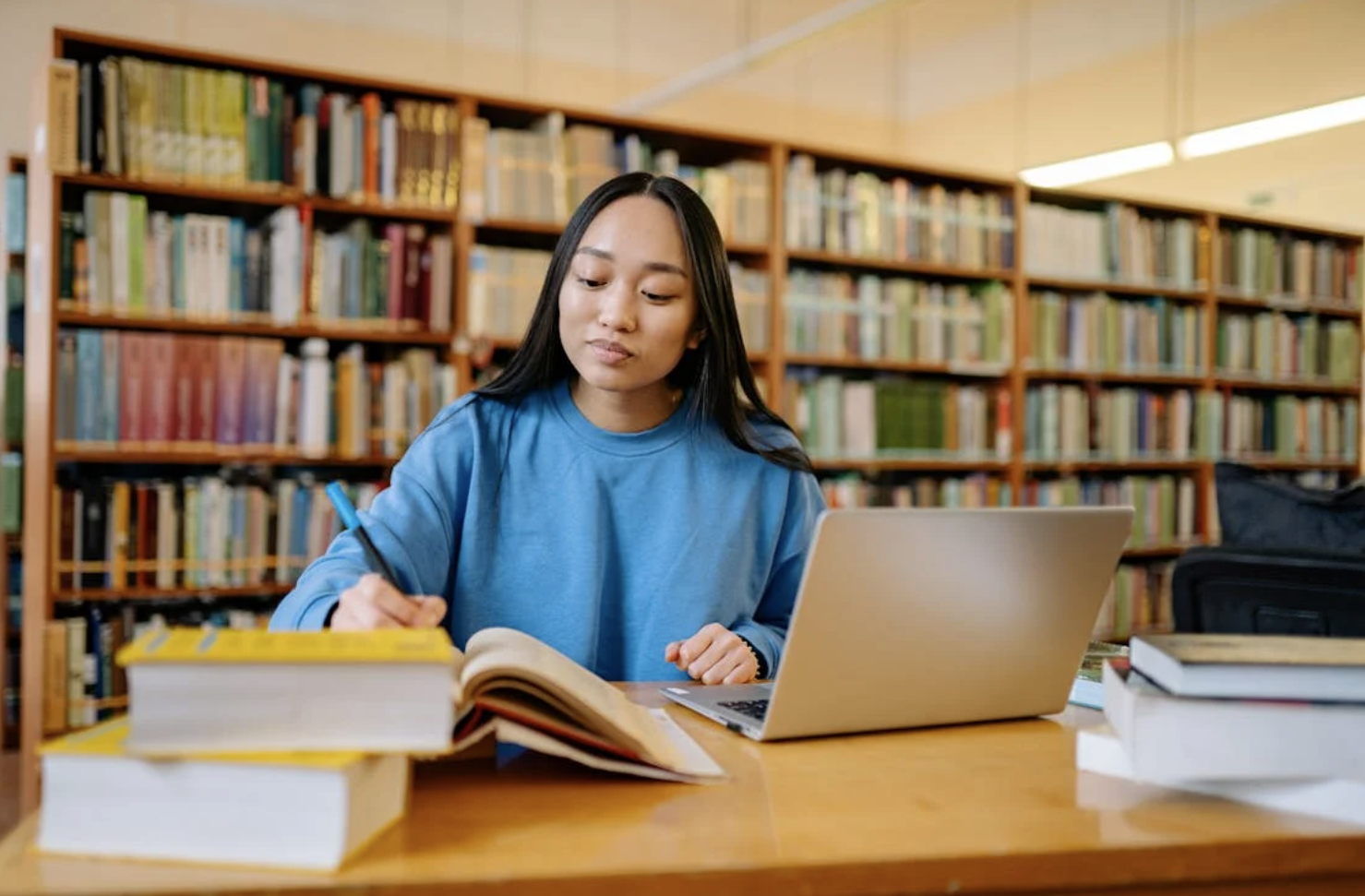 Student looks through a thick book and uses her laptop at a library