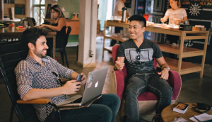 Two men, one on a laptop, smile and talk in a relaxed setting
