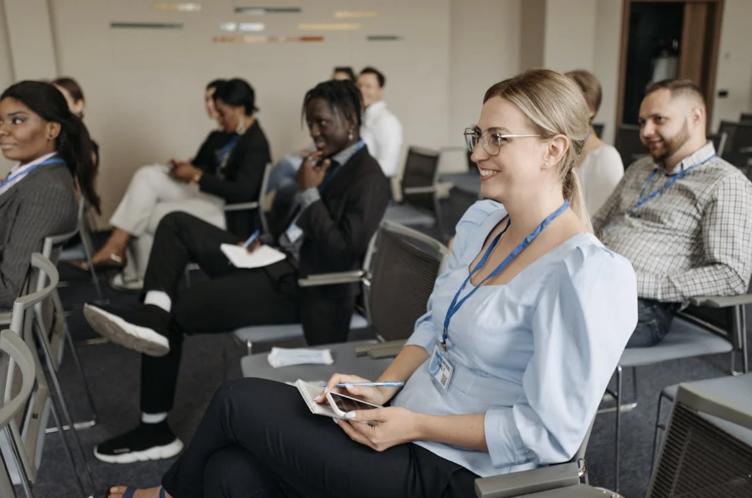 Woman in light blue shirt sits in rows of conference seating and smiles