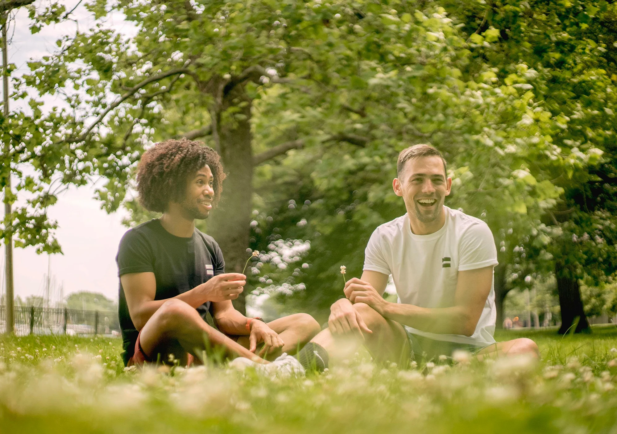 Two men sit in the grass, smiling and laughing, with trees behind them