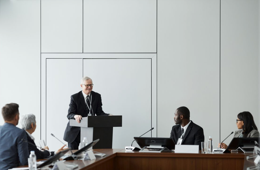 One man presents at podium while others sit near him at a large desk