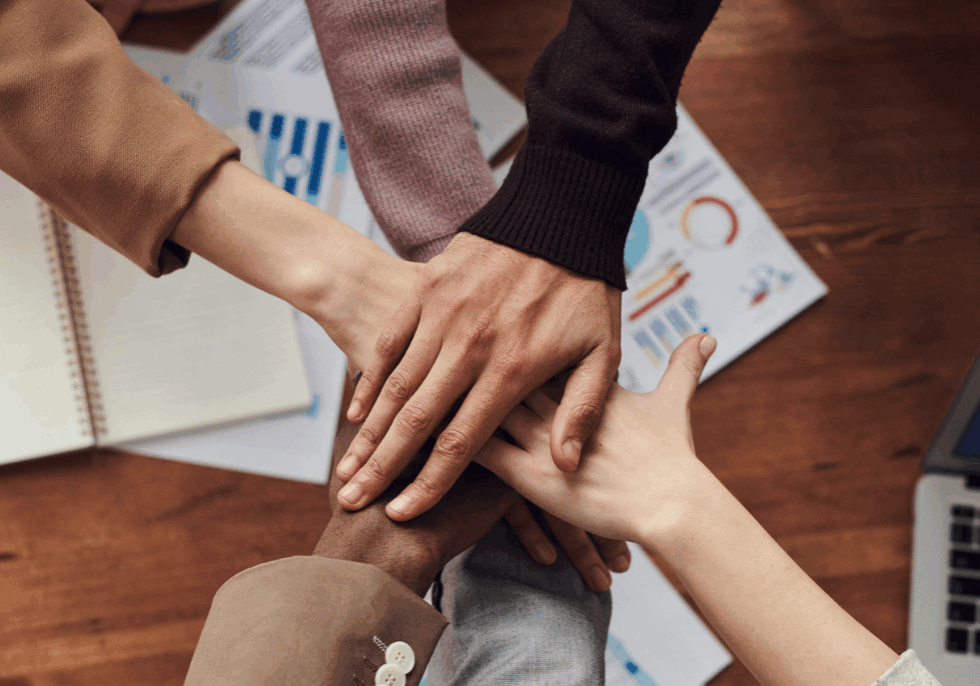 People put their hands together above a wooden table with data sheets and a laptop on it
